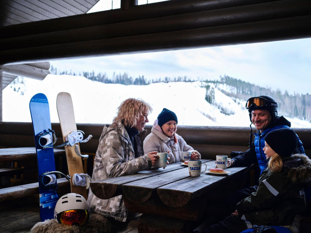 perhe rinneravintolassa tauolla talvivarusteissa, lumilaudat ja Himoksen laskettelurinne taustalla.