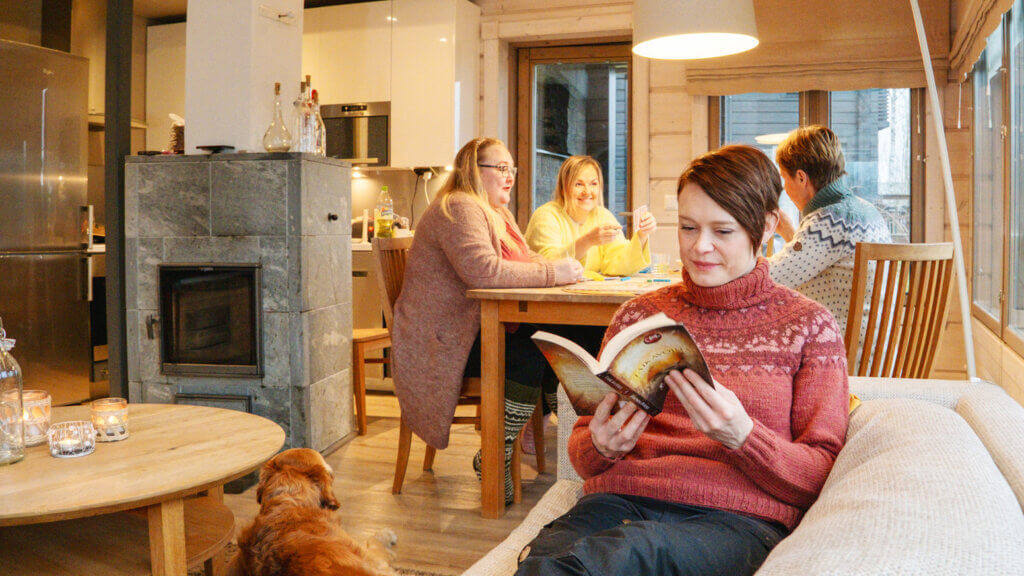 Mökin olohuoneessa yksi henkilö lukee kirjaa, koira makaa lattialla ja kolme ihmistä pelaa lautapeliä taustalla.