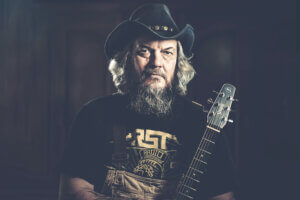 Cowboyhattu päässä oleva parrakas henkilö katsoo vakavana kameraan kitara kädessä.