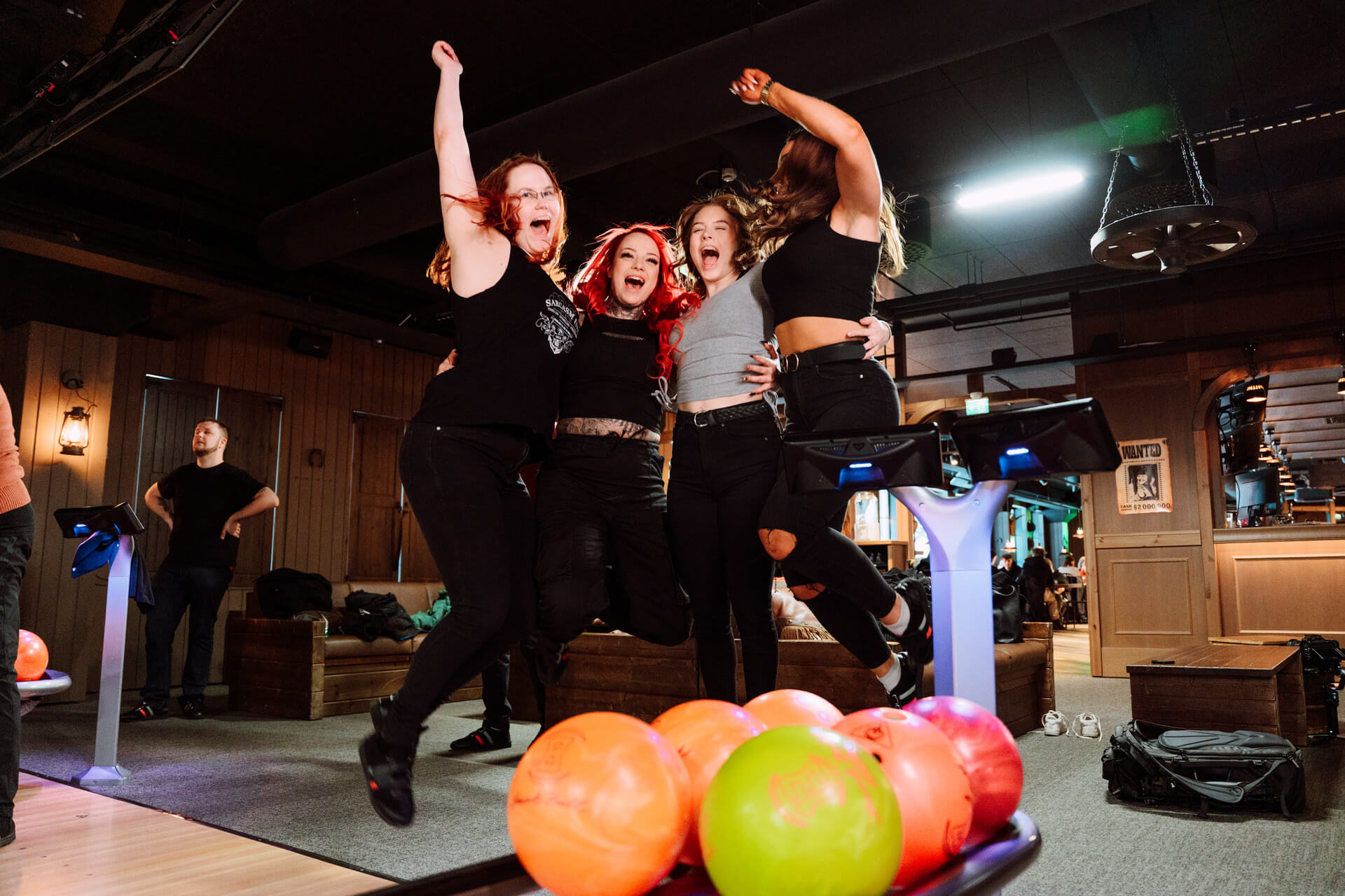 Neljä' henkilöä hypähtää ilmaan, edessä keilapalloja. Kuva Willi Länsi -huvikeskuksen keilahallista.