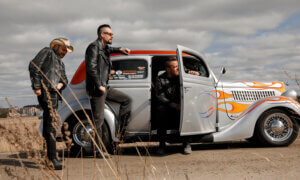 Kolme henkilöä katsoo oikealle kuvassa. Yhdellä on cowboyhatty, toinen norjaa autoon ja kolmas istuu autossa ja toinen etuovi on auki. Auto on hopeanvärinen, jossa on punainen katto ja keulassa on oransseja lieskoja. Auto näyttää vanhalta.