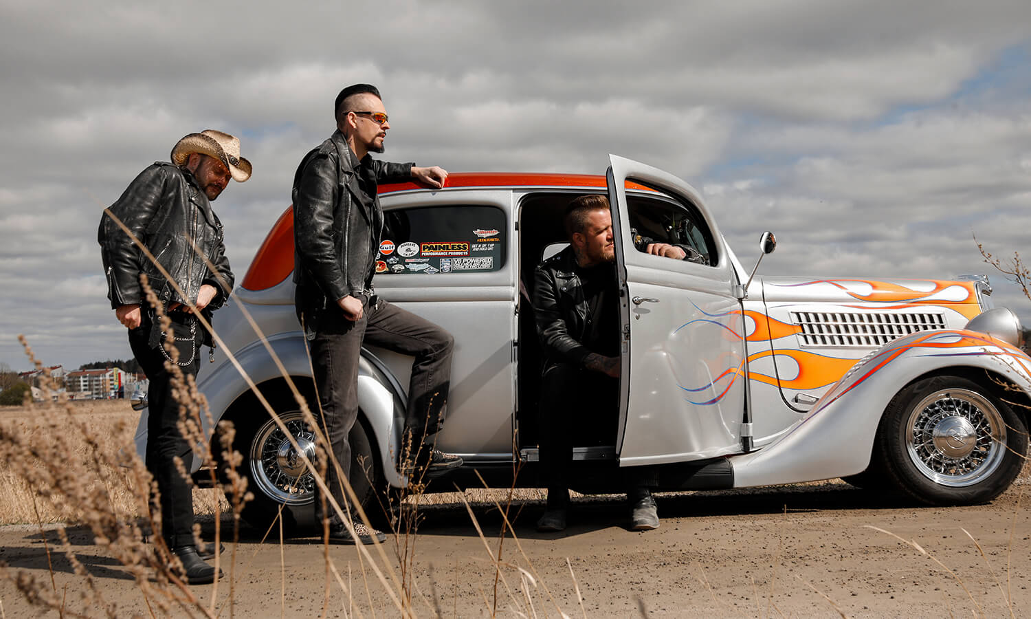 Kolme henkilöä katsoo oikealle kuvassa. Yhdellä on cowboyhatty, toinen norjaa autoon ja kolmas istuu autossa ja toinen etuovi on auki. Auto on hopeanvärinen, jossa on punainen katto ja keulassa on oransseja lieskoja. Auto näyttää vanhalta.