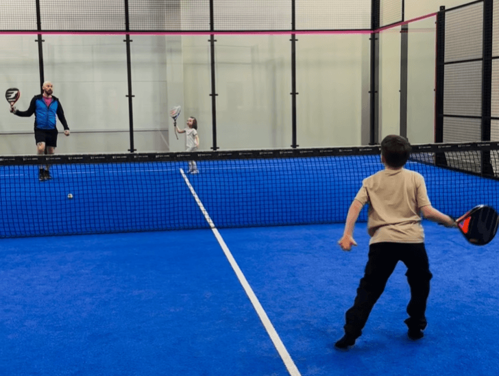 Kolme henkilöä, vanhempi ja kaksi nuorempaa, pelaavat padelia sinisellä pelialustalla sisähallissa.