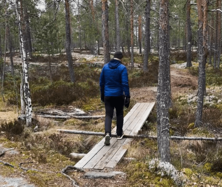 Henkilö kävelee selkä kameraan metsässä lankuista tehtyä siltaa pitkin Himoksella.