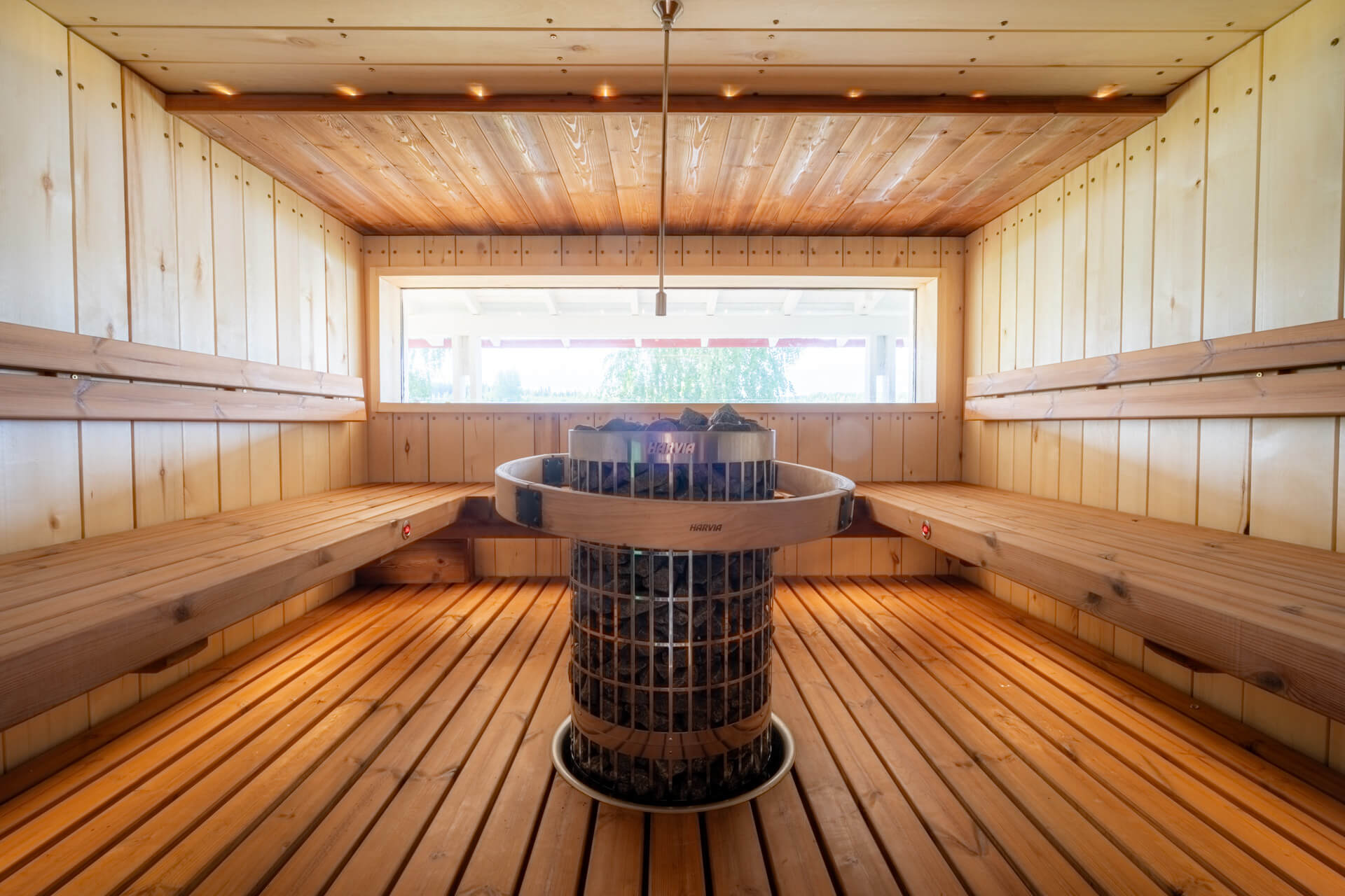 Puinen sauna, jonka keskellä iso harvia-merkkinen kiuas. Ikkuna, josta näkyy vihreää.