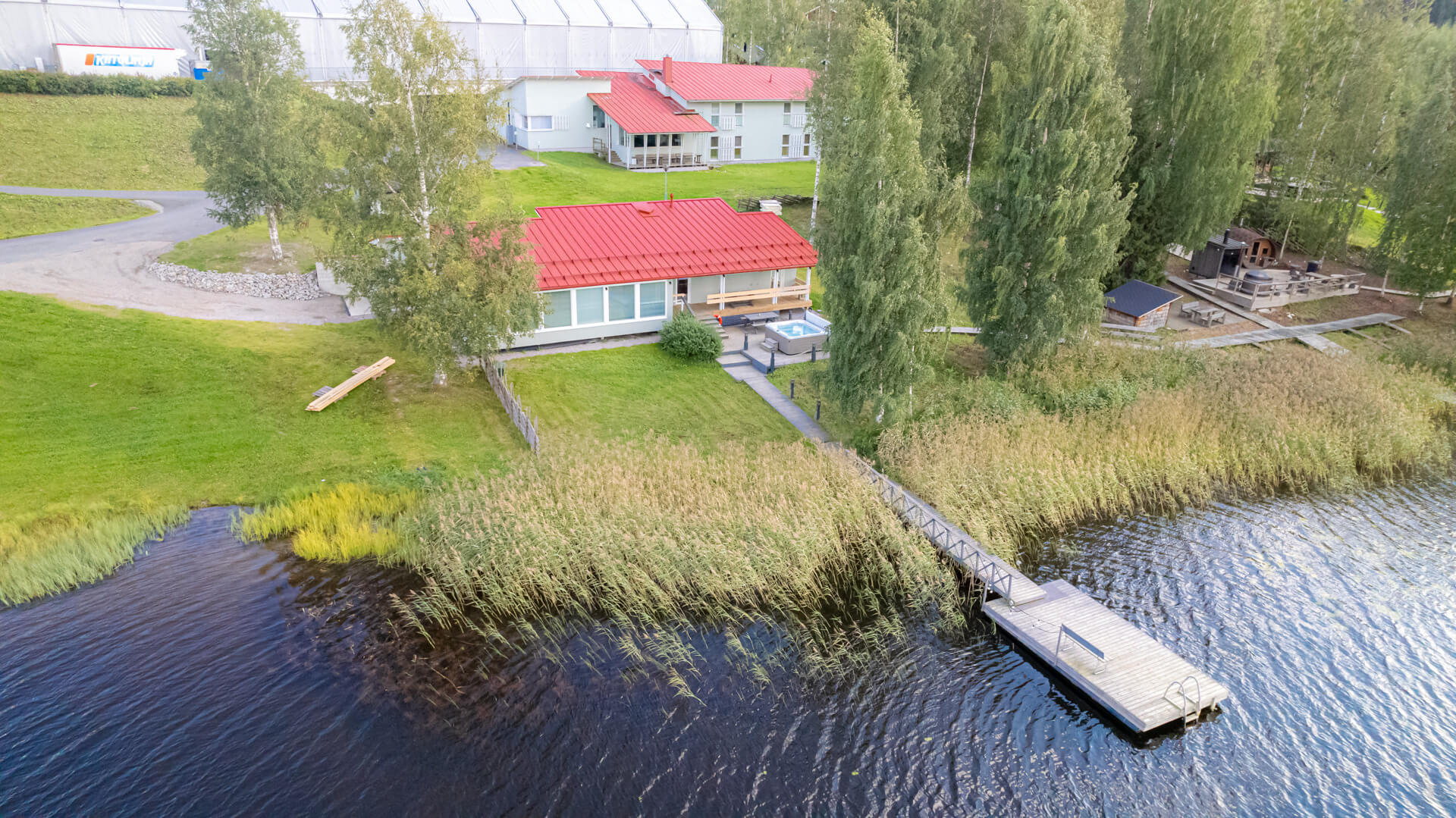 Himoksen Rantasauna kesällä. Pitkä laituri, luonnon kasvillisuutta ja punakattoinen rakennus, jossa isot ikkunat. Terassilla ulkoporeallas.