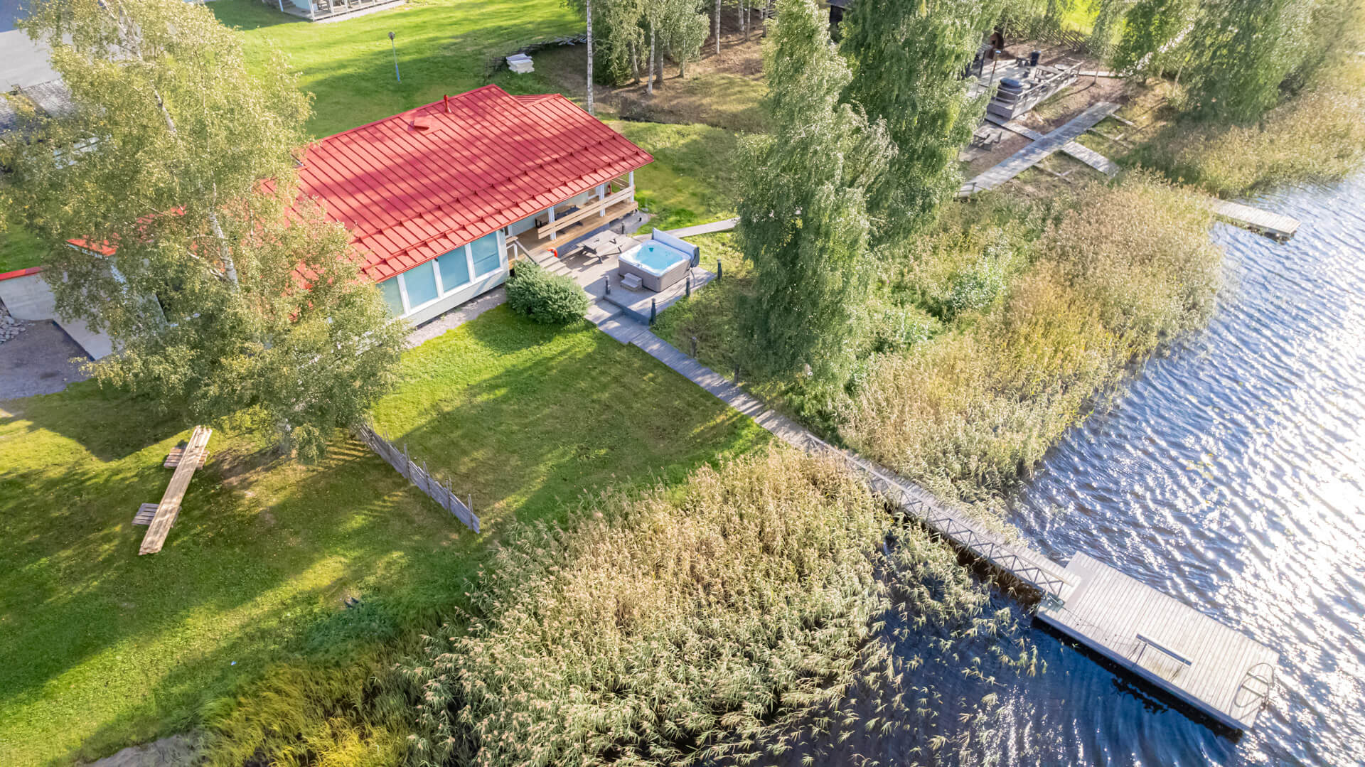 Ilmakuva Himoksen rantasaunasta. Pitkälaituri, vihreää nurmikkoa ja punakattoinen saunarakennus, jonka terassilla on poreallas. Kuvassa näkyy myös saunapihan toinen terassi sivulla.