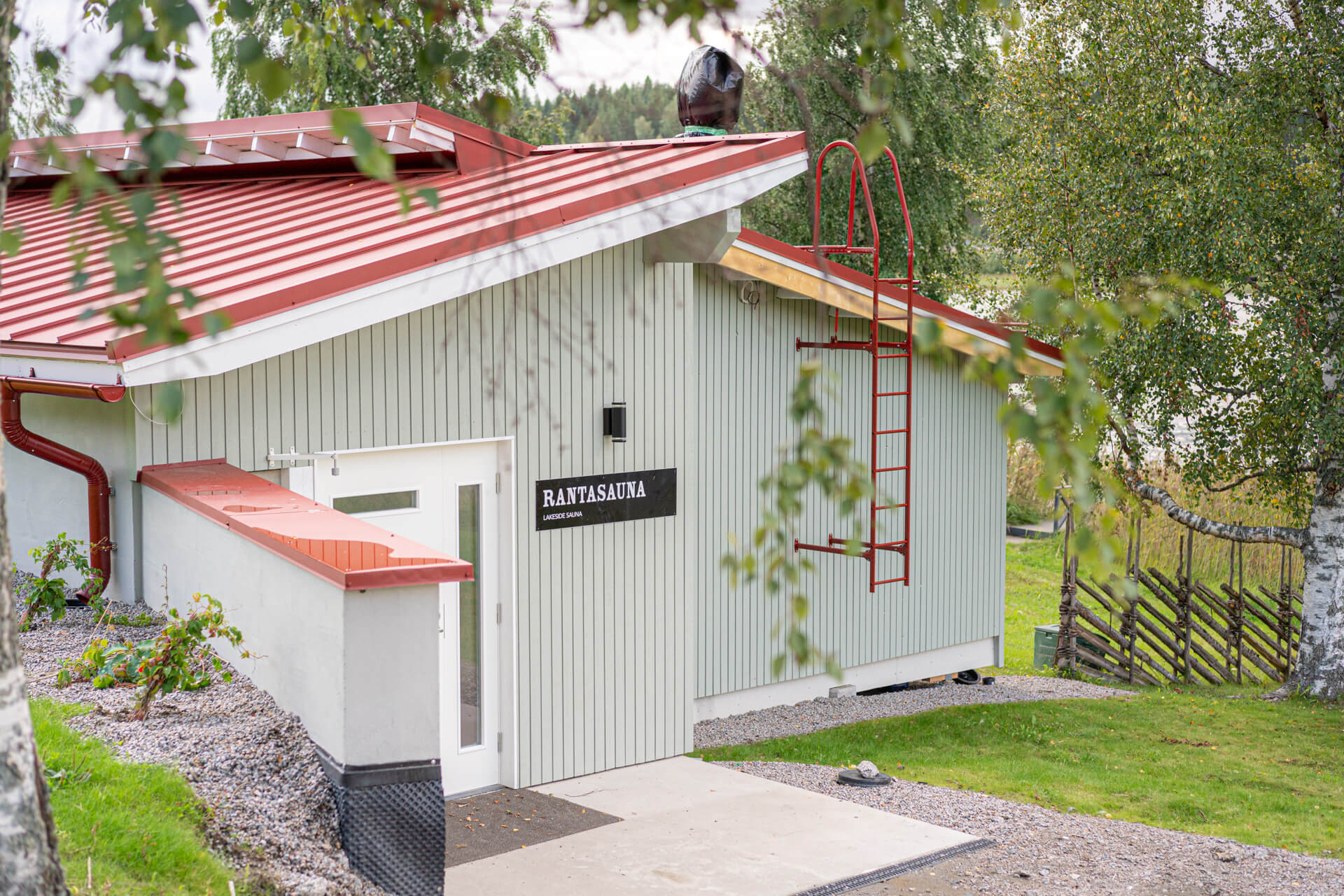 Kesäinen kuva HimosLomien Rantasaunasta. Vaalea rakennus, jossa punainen katto, kuvan sivussa riukuaita