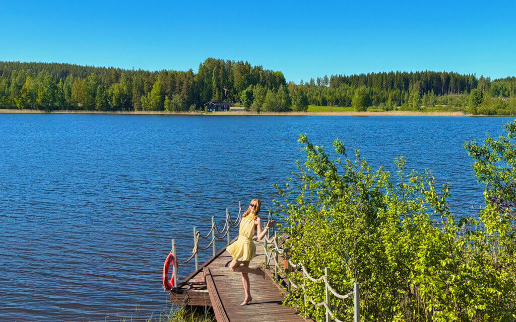 Kesäinen kuva, laiturilla seisoo henkilö mekossa, taustalla sininen järvi ja vihreää luontoa.