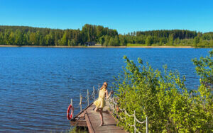 Kesäinen kuva, laiturilla seisoo henkilö mekossa, taustalla sininen järvi ja vihreää luontoa.