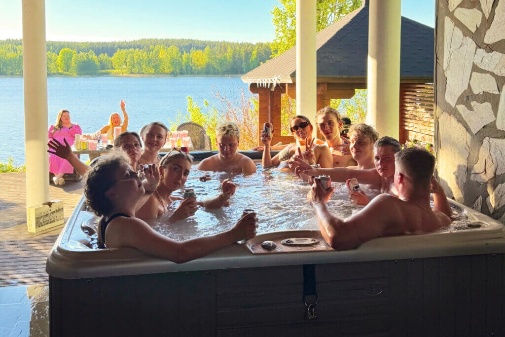 Iso joukko ihmisiä ulkoporealtaassa huvilan terassilla, takana näkyy järvi ja muutama ihminen sekä aurinkoa.