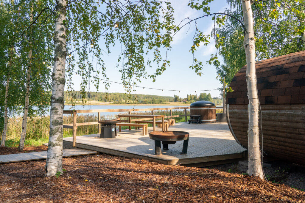 Himoksen Saunapiha kesällä takaapäin kuvattuna. Edessä hakkeesta tehty pohja, puinen iso terassi, jonka päällä näkyy tynnyrisauna. Kaksi isoa koivua joissa on runsaasti lehtiä.