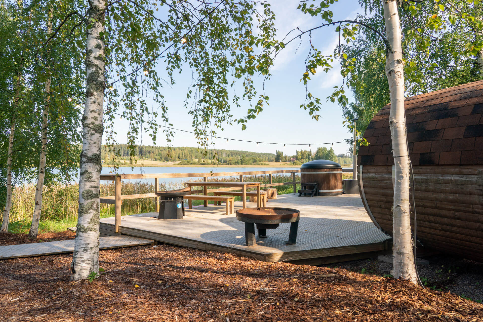 Himoksen Saunapiha kesällä takaapäin kuvattuna. Edessä hakkeesta tehty pohja, puinen iso terassi, jonka päällä näkyy tynnyrisauna. Kaksi isoa koivua joissa on runsaasti lehtiä.