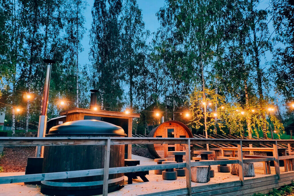 Iltavalaistuksessa, paljon pieniä keltaisena hehkuvana lamppuja puinen iso saunaterassi, jonka päällä näkyy palju ja taustalla pieni sauna. Kuva Himoksen Saunapihasta.
