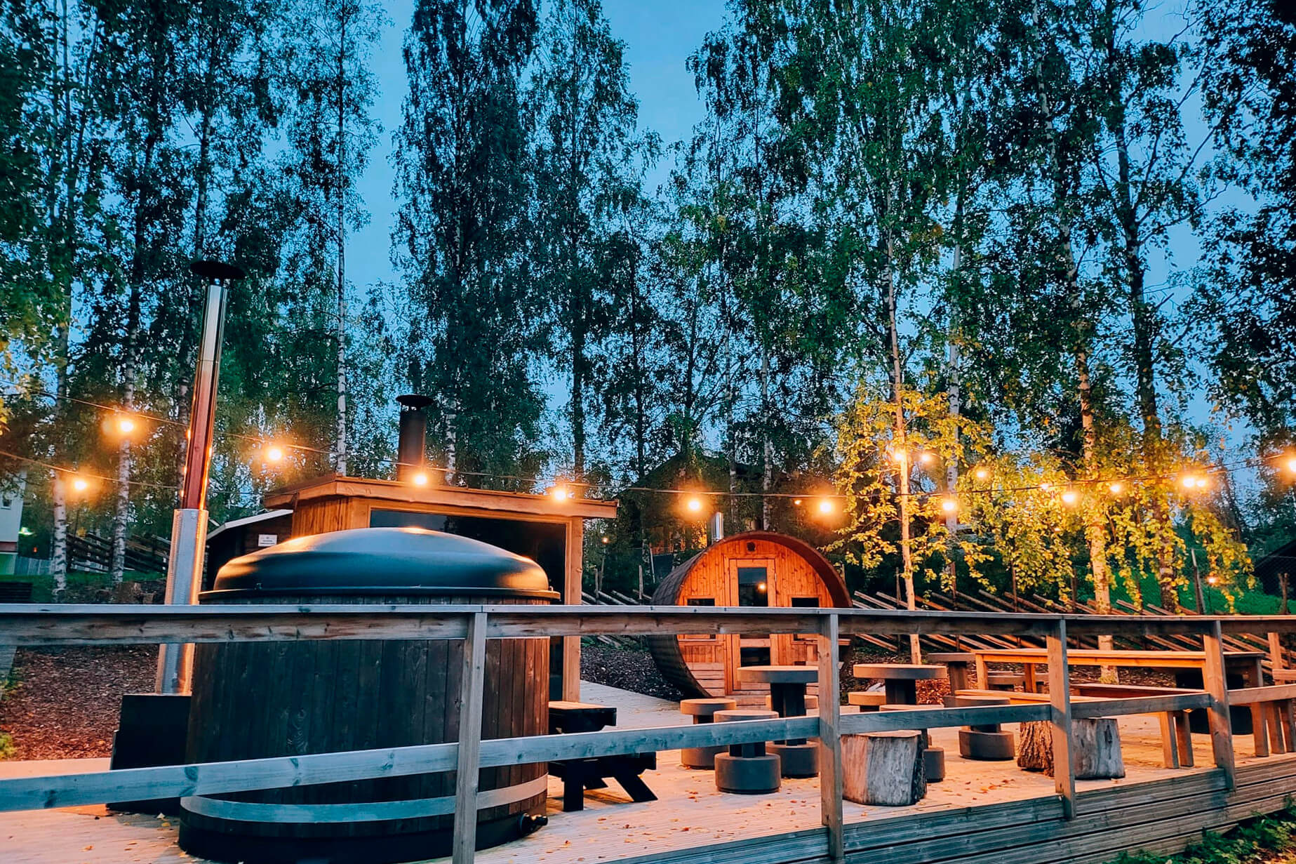 Iltavalaistuksessa, paljon pieniä keltaisena hehkuvana lamppuja puinen iso saunaterassi, jonka päällä näkyy palju ja taustalla pieni sauna. Kuva Himoksen Saunapihasta.