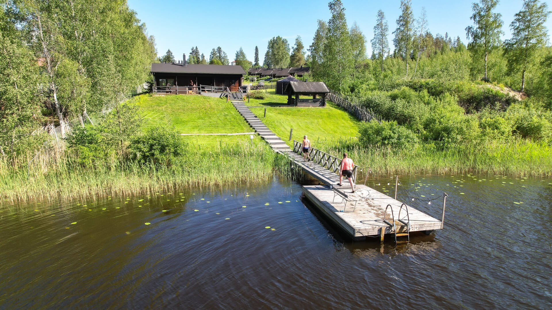 Savusauna Himoksella. Kesäinen kuva järveltä, pitkällä laiturilla kävelee henkilö kohti savusaunaa, joka on kuvassa. Paljon vihreää, on kesä.