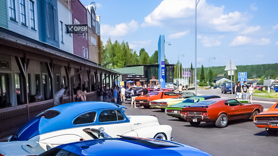 Paljon amerikkalaisia autoja aurinkoisessa säässä parkkeerattuna Willi Länsi -huvikeskuksen eteen Himoksella. Taivas on sininen ja kuvassa näkyy eri värisiä ja vuosimallin sekä merkin classic-autoja.