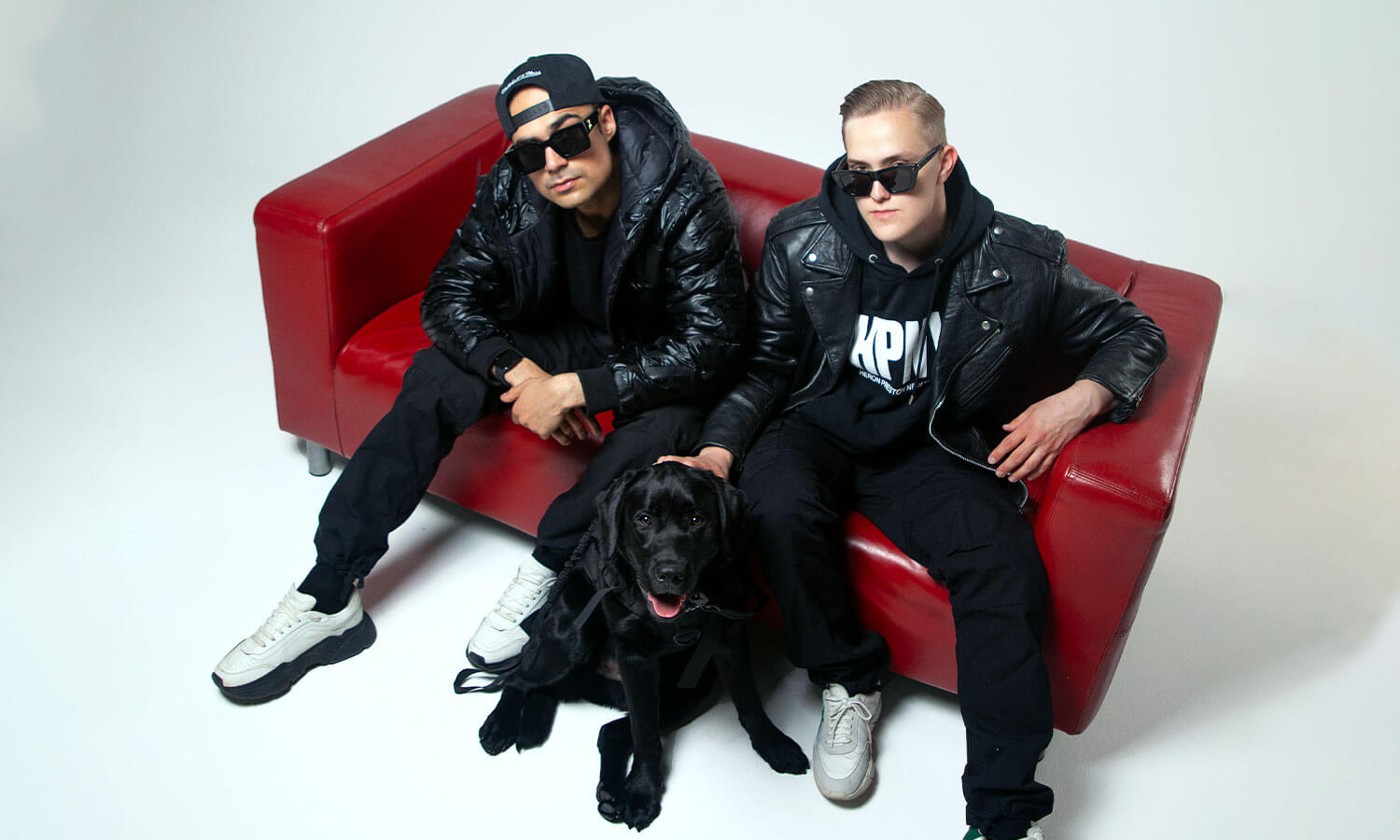 Dog Hustlers -duo istuvat punaisella sohvalla mustissa vaatteissa. Edessään musta labradorinnoutaja, joka istuu sohvan edessä.