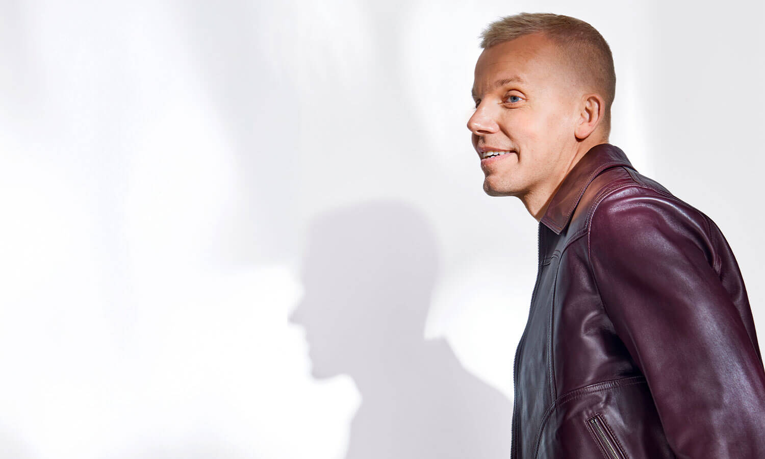 Vaalea kuva, jossa artisti Elastinen on oikeassa reunassa, kuvassa näkyy myös Elastisen varjo. Artisti katsoo vasemmalle sivuun hieman hymyillen, hänellä on tumma takki.