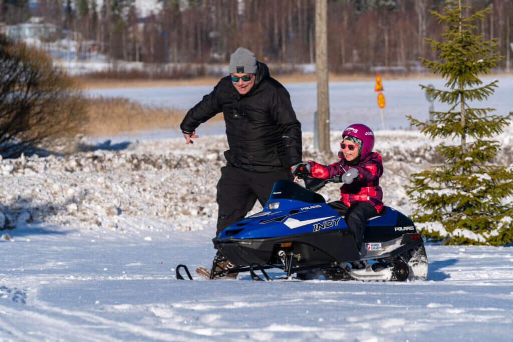 Lapsi moottorikelkan kyydissä Himoksella. Aikuinen juoksee vieressä. Lapsi nauraa suu auki.