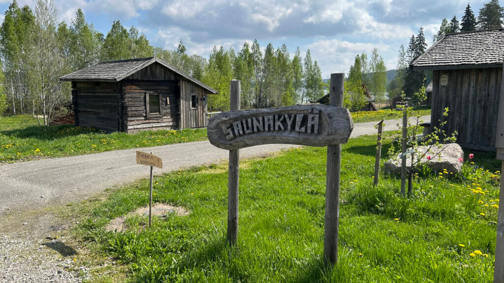 Kyltti Jämsän Saunakylästä, tehty puusta. Taustalla näkyy savusaunoja kesäisessä maisemassa, Paljon vihreää.