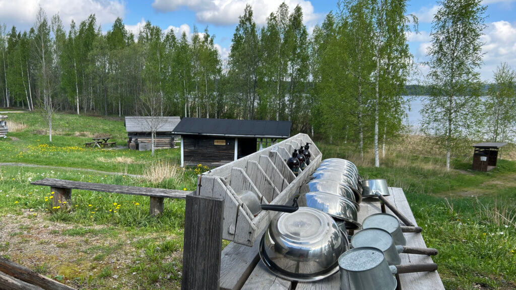 Kesä Jämsän Saunakylässä. Edessä metallisia pesuvateja ja kiuluja, taustalla koivujen takana näkyy Päijänne.