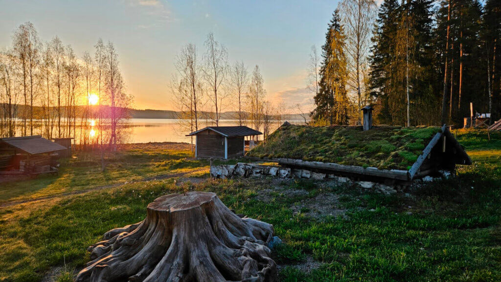Jämsän Saunakylä iltavalaistuksessa, Taustalla aurinko laskee Päijänteen taakse, edessä iso kanto ja sen takana matala sammalkattoinen savusauna. Kesäinen kuva iltavalaistuksessa.