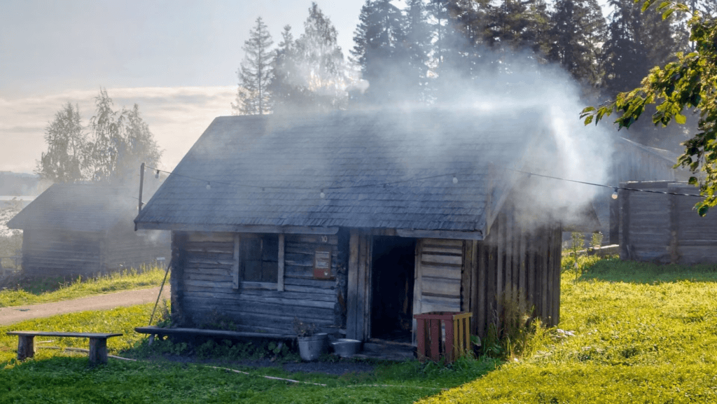 Kesä ja Vallius-niminen savusauna Jämsän Saunakylässä. Savua tulee savusaunan hirsienkin läi, aurinkopaistaa.