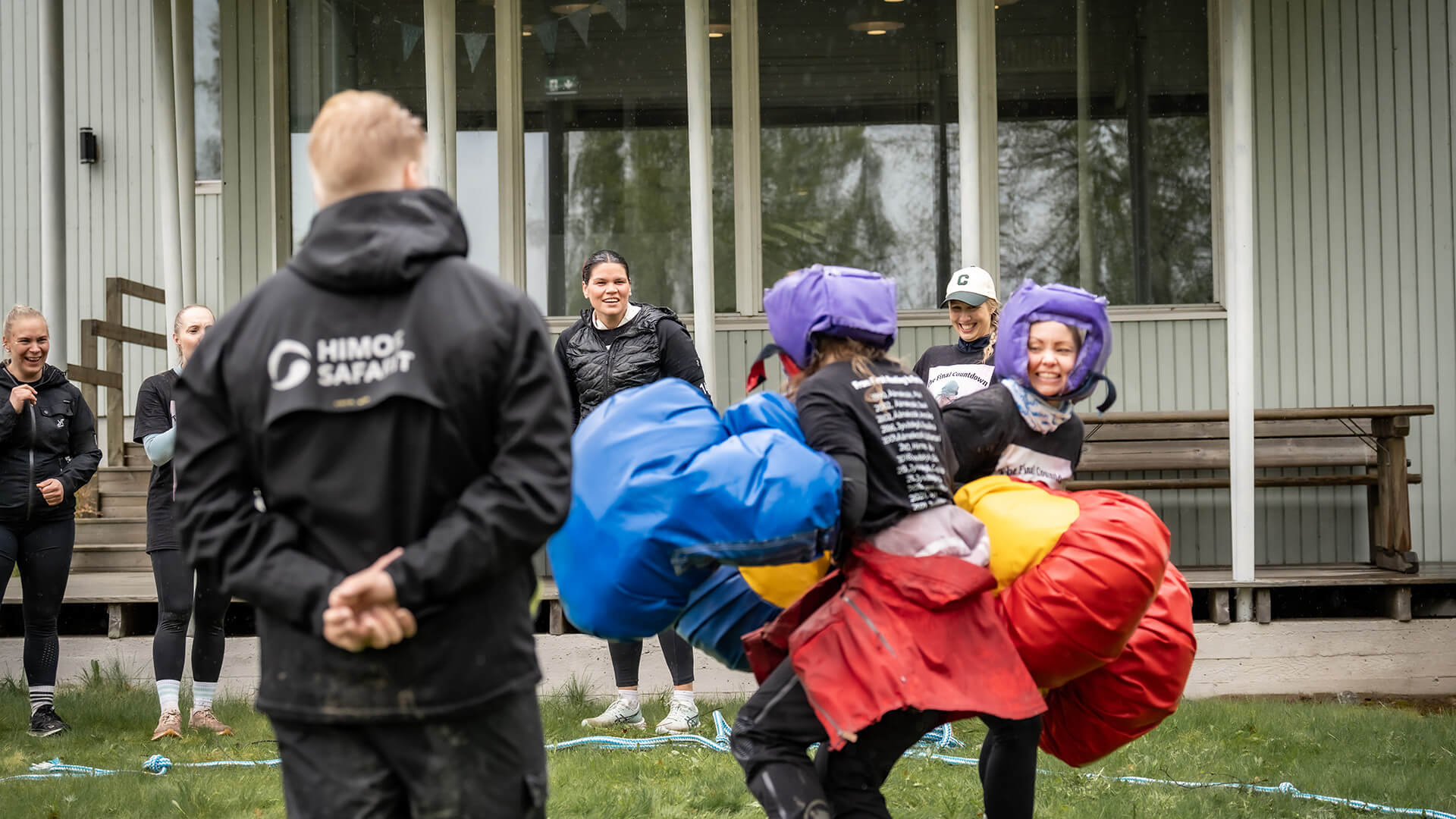 Kaksi henkilöä nyrkkeilee pehmonyrkein ja hymyilevät. Taustalla ihmisiä kannustamassa, yksi etualalla selin kamerana, mustan takin selässä lukee Himos Safarit.