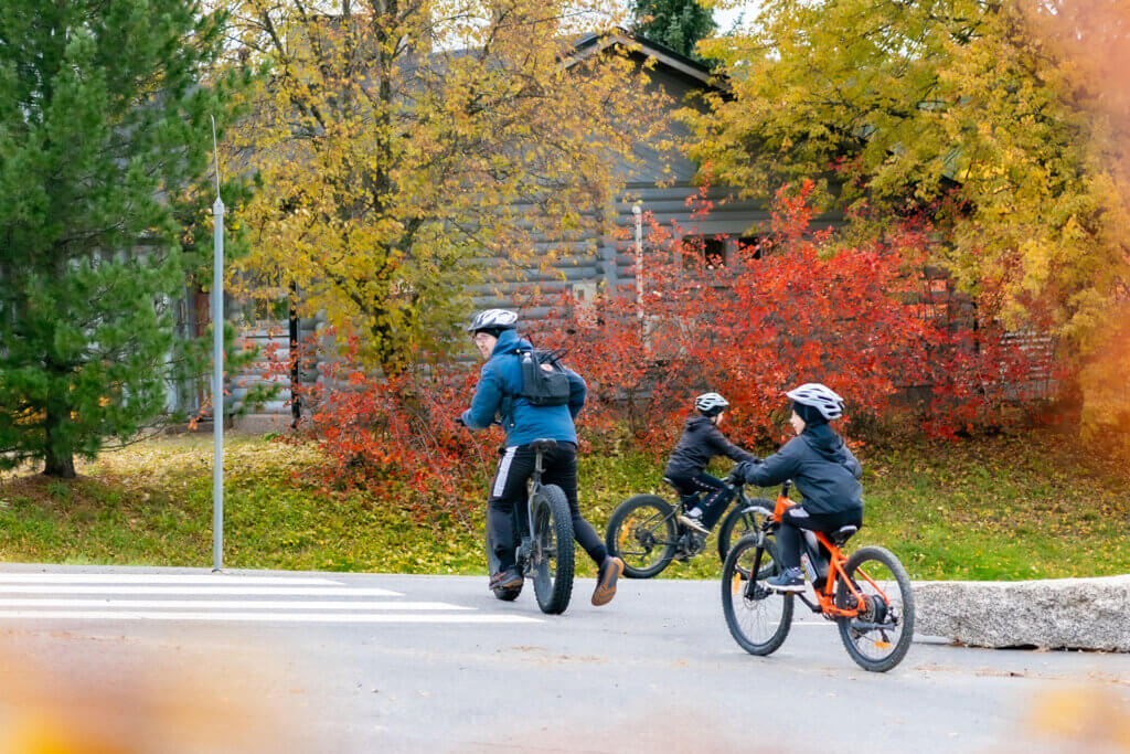 Syksyn värikkäässä maisemassa aikuinen ja kaksi lasta ylittämässä tietä sähköavusteisilla pyörillä.