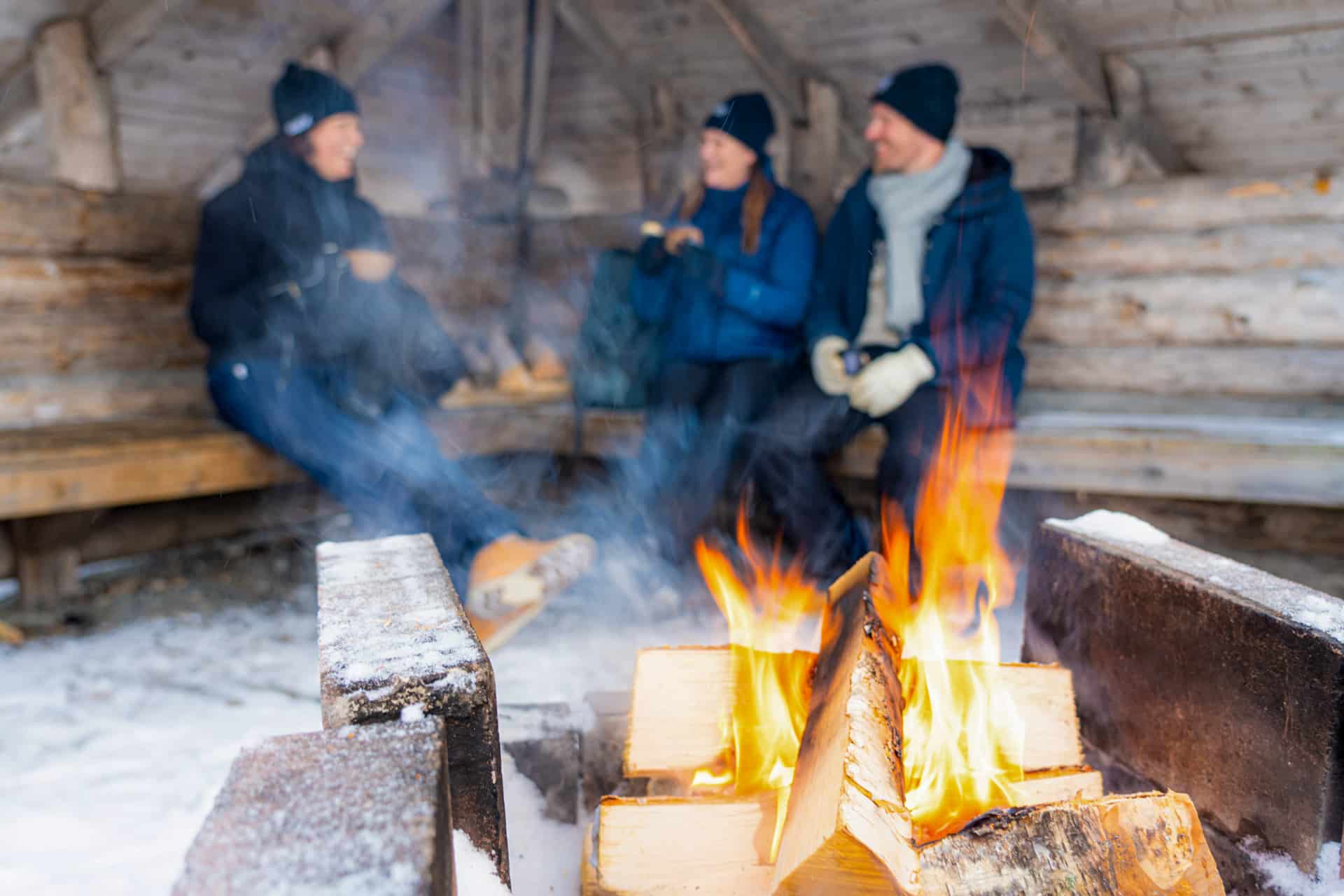 kolme ihmistä hymyilee taustalla nuotion ääressä.