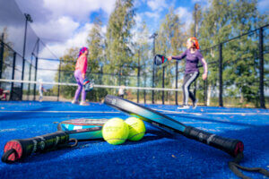 Kaksi ihmistä padelkentällä taustalla, etualalla mailat ja pallot.