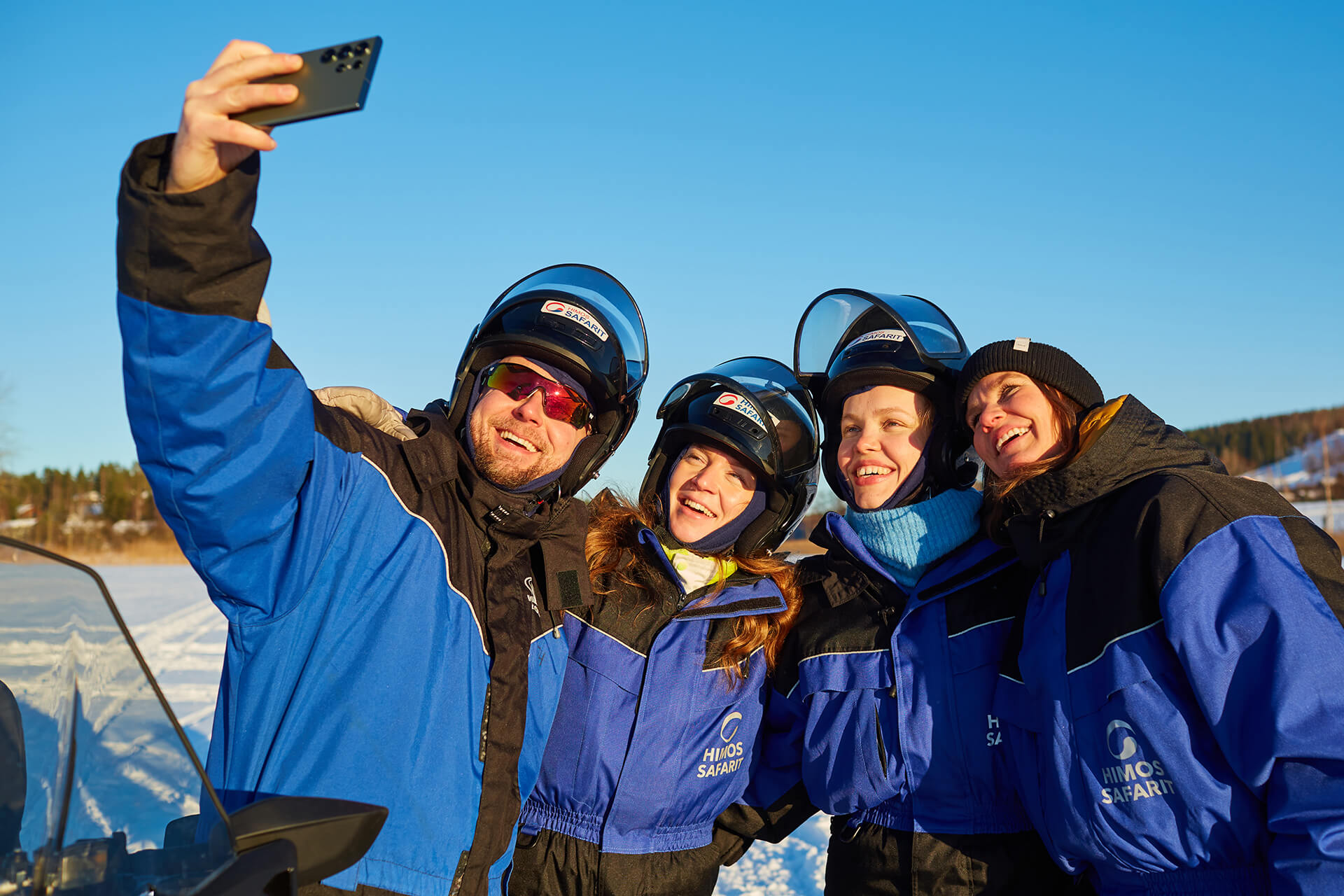 Moottorikelkkaporukka kuvaa selfietä ja nauttii aurinkoisesta talvesta.